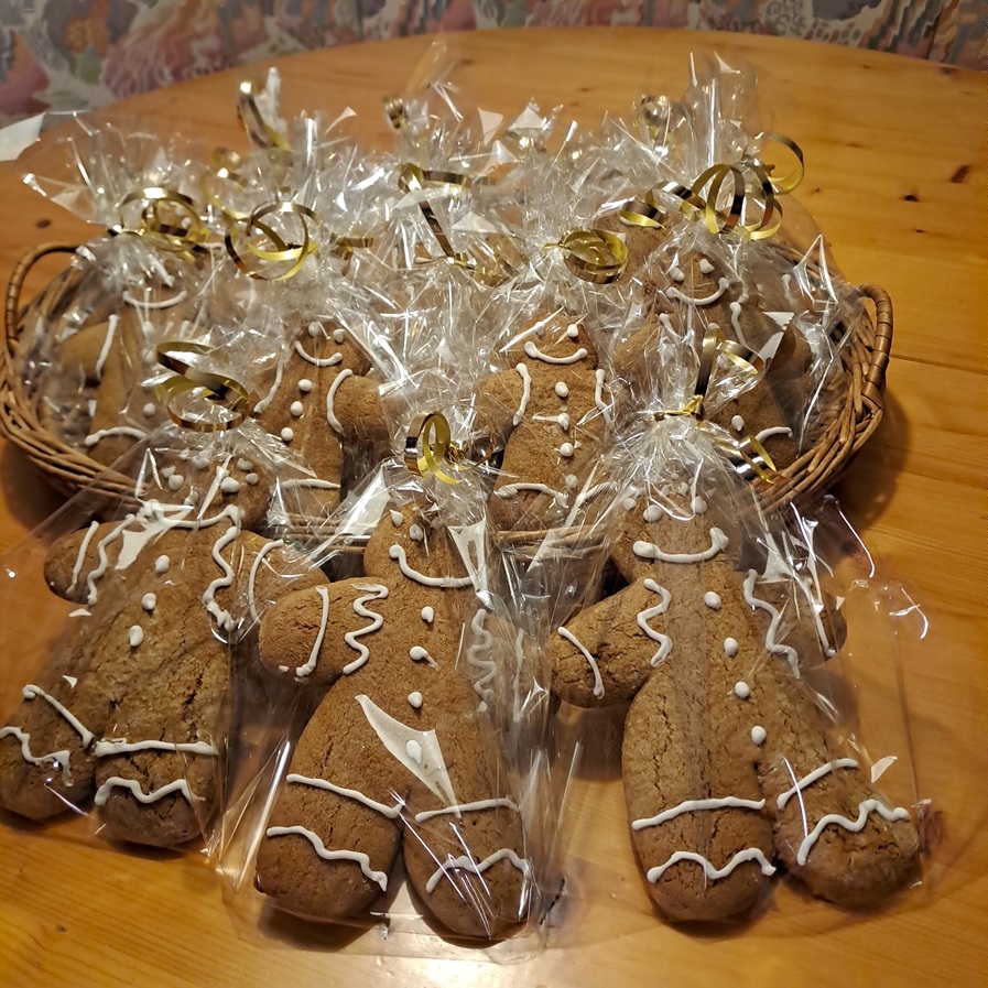 Ein Korb voller Lebkuchen in Menschenform, jeder mit weißem Zuckerguss verziert und einzeln in transparente Plastikfolie eingewickelt und mit goldenen Schnüren zusammengebunden. Der Korb steht auf einem Holztisch.