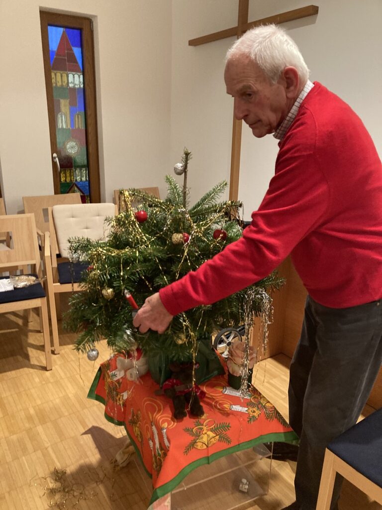 Ein älterer Mann in einem roten Pullover schmückt einen kleinen Weihnachtsbaum mit Lametta und Ornamenten. Der Baum steht auf einem mit einem festlichen Tuch bedeckten Tisch. Im Hintergrund sind ein Buntglasfenster und Holzstühle zu sehen.