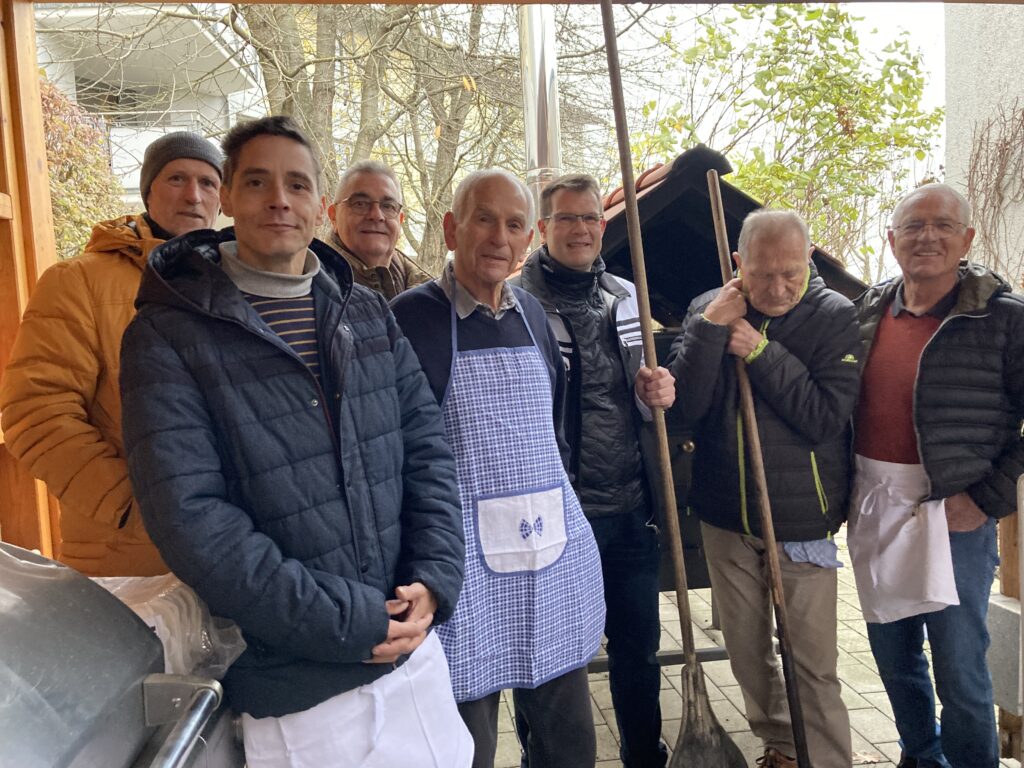 Eine Gruppe von sieben Männern steht im Freien zusammen. Sie tragen Freizeit- und Arbeitskleidung. Einige halten Werkzeuge oder tragen Schürzen, was darauf hindeutet, dass sie einer Aktivität nachgehen. Im Hintergrund sind Bäume und ein Gebäude zu sehen.