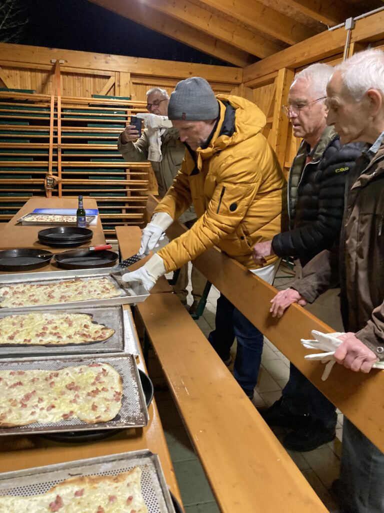 Menschen versammeln sich drinnen um einen langen Holztisch mit mehreren Tabletts voller Essen. Eine Person in einer gelben Jacke stellt ein Tablett auf den Tisch. Andere schauen zu. Im Hintergrund sind Klapptische und Holzvertäfelungen zu sehen.