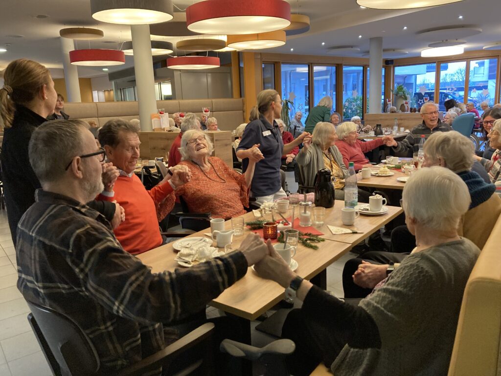 Eine Gruppe älterer Menschen sitzt in einem hellen, geräumigen Raum um einen Tisch und genießt gemeinsam eine Mahlzeit. Sie halten sich an den Händen und lächeln, wodurch eine warme, freudige Atmosphäre entsteht. Mitarbeiter helfen und unterhalten sich mit der Gruppe.