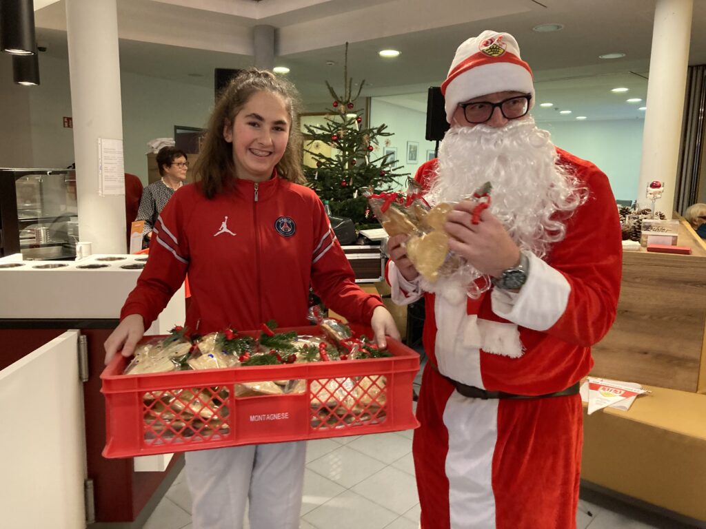 Eine als Weihnachtsmann verkleidete Person und eine andere Person in einer roten Jacke lächeln, während sie eine Kiste mit Geschenktüten halten. Im Hintergrund steht ein geschmückter Weihnachtsbaum, der im Haus eine festliche Atmosphäre schafft.