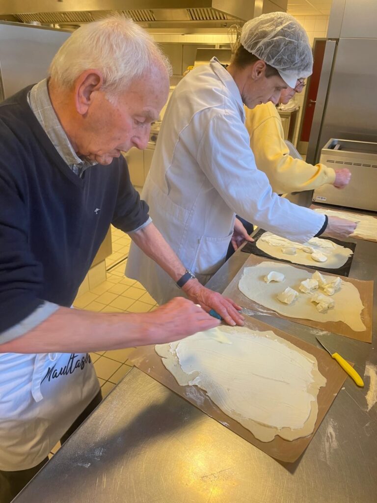 Ein älterer Mann und zwei Personen in weißen Laborkitteln verteilen in einer Großküche cremigen Käse auf großen Teigplatten.