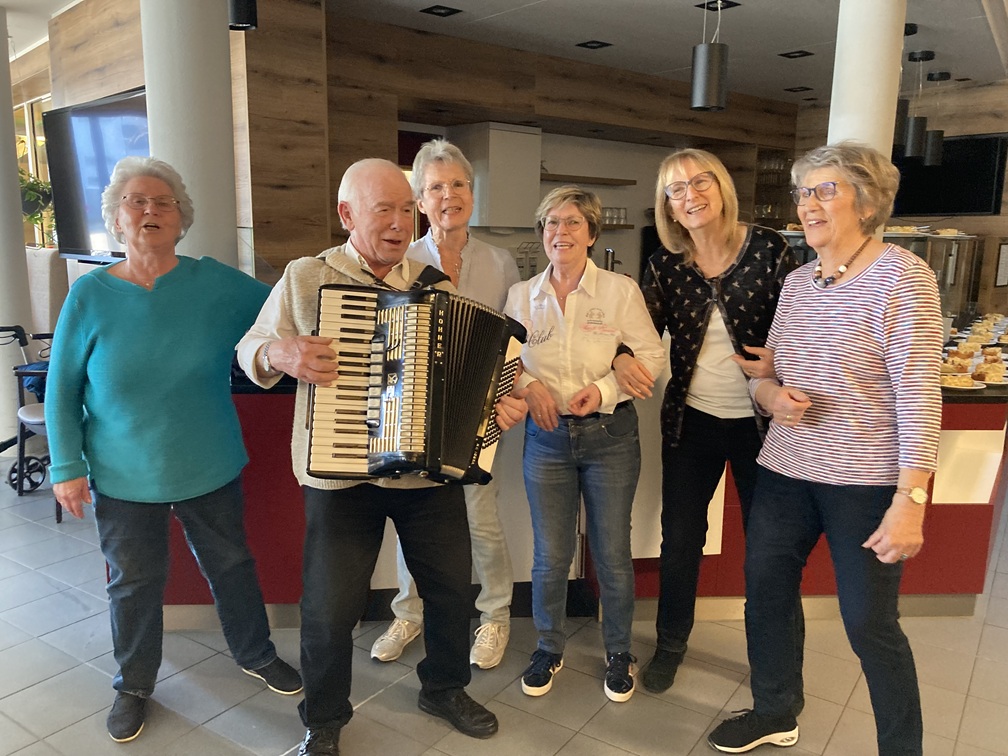 Eine Gruppe von sechs älteren Erwachsenen steht drinnen auf einem gefliesten Boden. Ein Mann in der Mitte spielt Akkordeon, während die fünf Frauen um ihn herum lächeln und miteinander reden. Im Hintergrund sind Holzwände und ein Küchenbereich zu sehen.