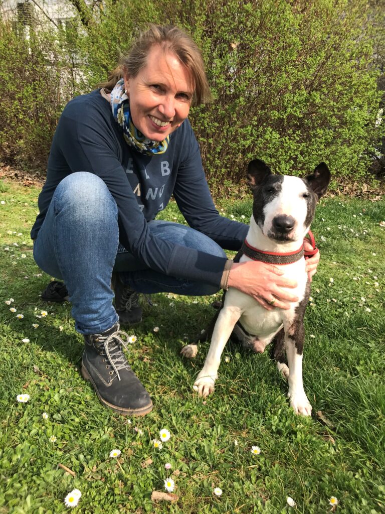 Eine Frau hockt im Gras und lächelt in die Kamera, während sie einen schwarz-weißen Hund mit rotem Geschirr hält. Sie befinden sich im Freien, im Hintergrund sind Grünflächen und kleine weiße Blumen zu sehen.