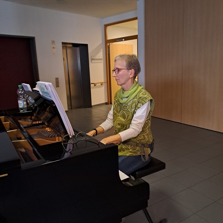 Eine ältere Frau mit Brille und grün gemusterter Weste spielt drinnen auf einem schwarzen Flügel und liest Noten. Sie sitzt auf einer schwarzen Klavierbank in einem modernen, gut beleuchteten Raum mit gefliestem Boden.