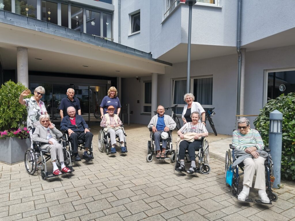 Eine Gruppe älterer Menschen, einige in Rollstühlen, posiert und lächelt vor einem Wohnhaus, hinter ihnen stehen zwei Frauen. Eine Frau winkt in die Kamera. Die Gruppe ist auf einer gepflasterten Fläche in der Nähe einer Grünanlage versammelt.