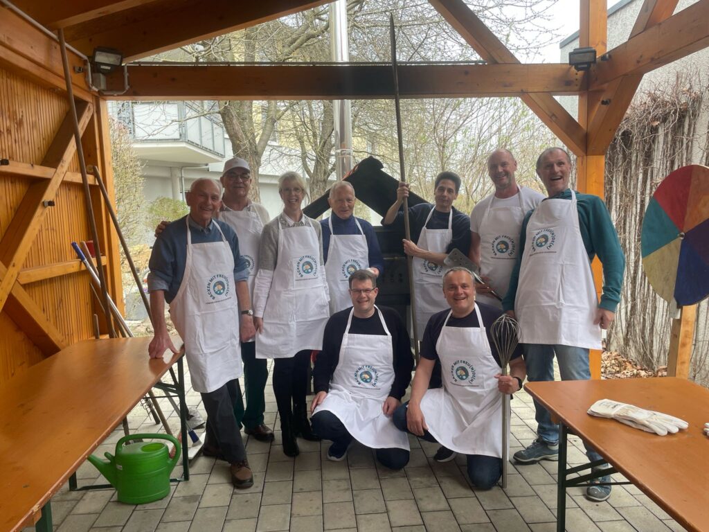 Eine Gruppe von neun Erwachsenen mit weißen Schürzen posiert und lächelt unter einem hölzernen Vordach. Einige halten Küchenutensilien in der Hand. Die Szene scheint im Freien zu spielen, mit Tischen, einer Gießkanne und Bäumen im Hintergrund.