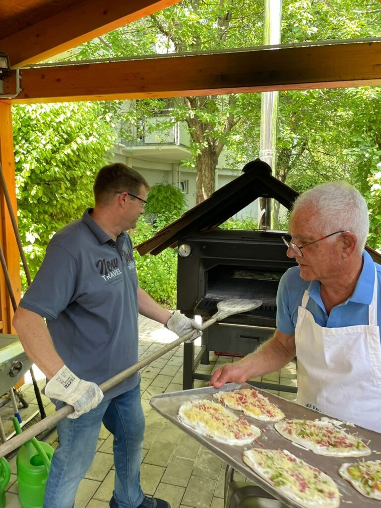 Zwei Männer bereiten im Freien Pizzen zu; einer schiebt eine Pizza in einen Holzofen, während der andere ein Tablett mit rohen Pizzen hält. Bäume und Grünflächen umgeben den überdachten Terrassenbereich.