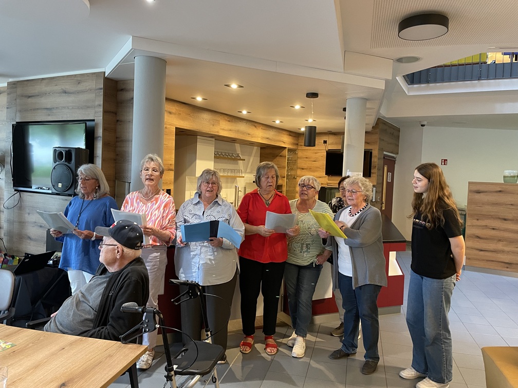 Eine Gruppe von sieben älteren Frauen steht drinnen und singt von Liederblättern, während zwei sitzende Personen zuhören. Der Raum ist modern mit Holzakzenten und einer entspannten Atmosphäre.