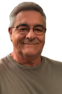 Ein lächelnder älterer Mann mit grauem Haar und Brille, der ein graues T-Shirt trägt, posiert vor einem schlichten weißen Hintergrund.