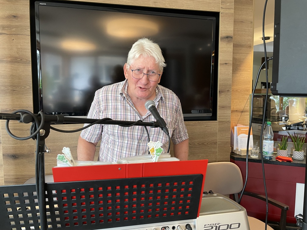 Ein älterer Mann mit weißem Haar und Brille steht hinter einem Keyboard und singt in ein Mikrofon. Er befindet sich in einem Raum mit einem großen Flachbildfernseher an der Wand hinter ihm und Notenblättern vor ihm.