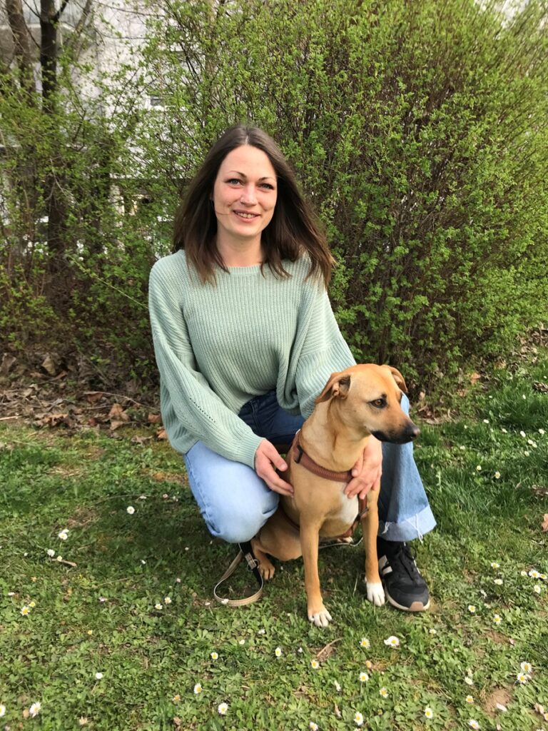 Eine Frau mit langen braunen Haaren, die einen grünen Pullover und Jeans trägt, kniet im Gras neben einem hellbraunen Hund mit einem roten Geschirr. Sie befinden sich im Freien mit grünen Büschen und Bäumen im Hintergrund.