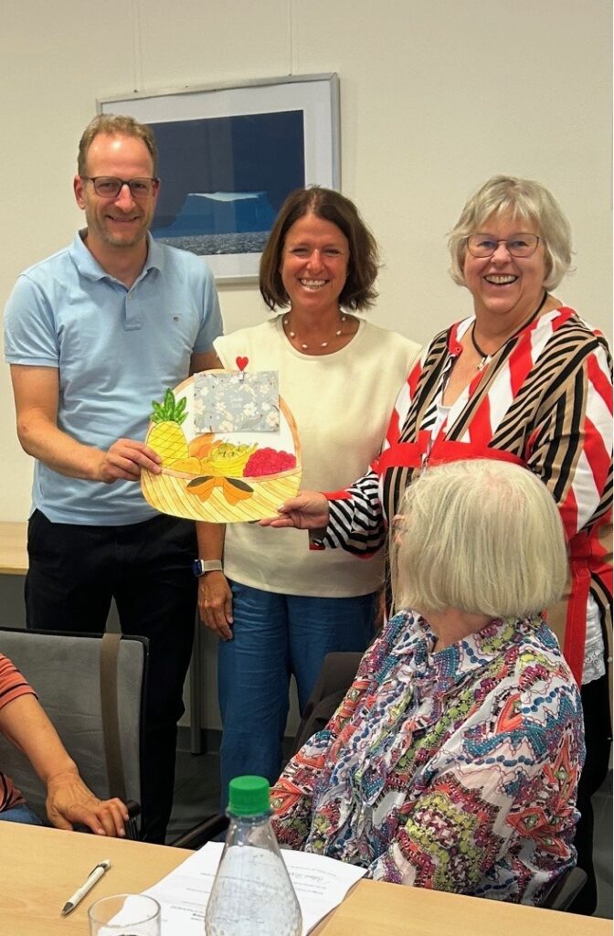 Drei lächelnde Erwachsene stehen und halten eine bunte, handgezeichnete Karte mit Obst darauf, während eine andere sitzende Frau in gemusterter Kleidung zusieht. Auf dem Tisch vor ihnen liegen Papiere und eine Wasserflasche.