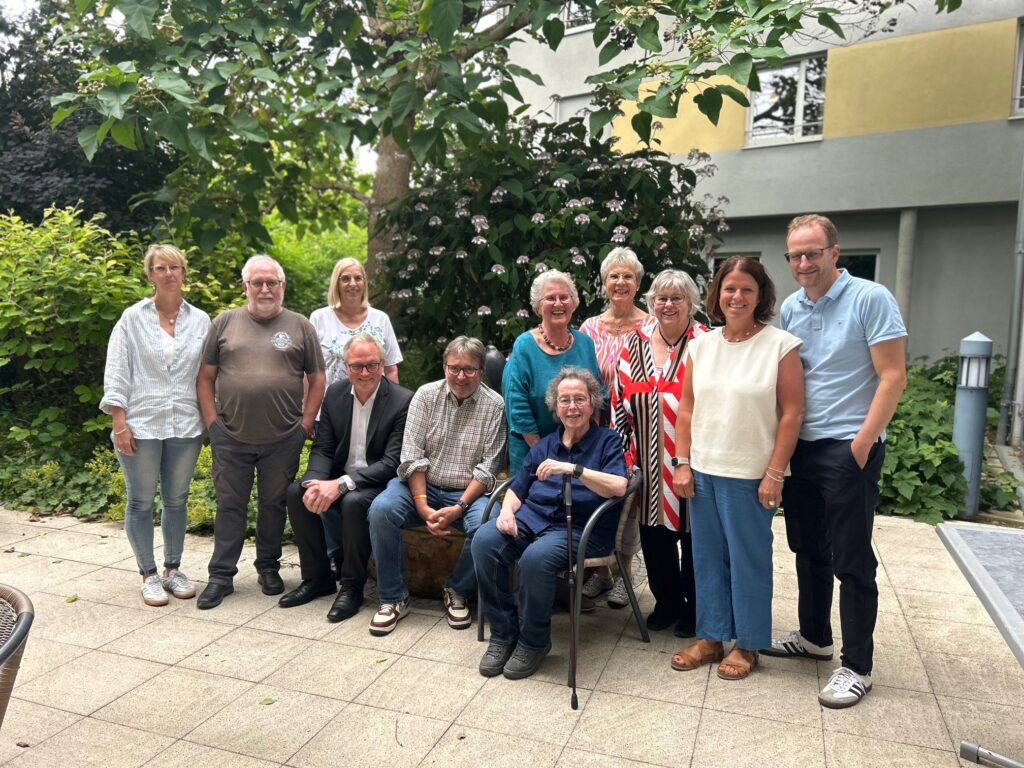 Eine Gruppe von zwölf Erwachsenen, teils stehend, teils sitzend, posiert gemeinsam auf einer Terrasse vor einem Gebäude mit viel Grün. Die meisten lächeln und sind leger gekleidet.