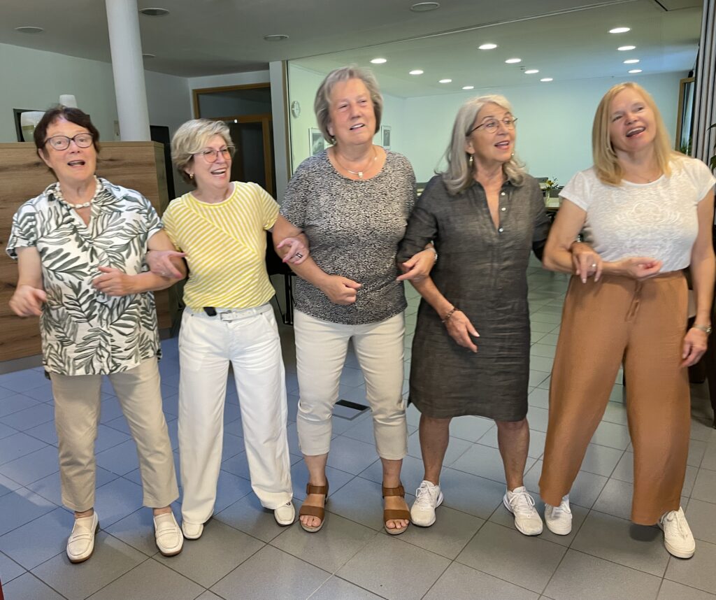 Fünf ältere Frauen stehen in einem Innenraum in einer Reihe, lächeln und verschränken die Arme, während sie miteinander singen oder sich unterhalten. Sie sind leger gekleidet und wirken fröhlich und entspannt in einem hellen, modernen Raum.