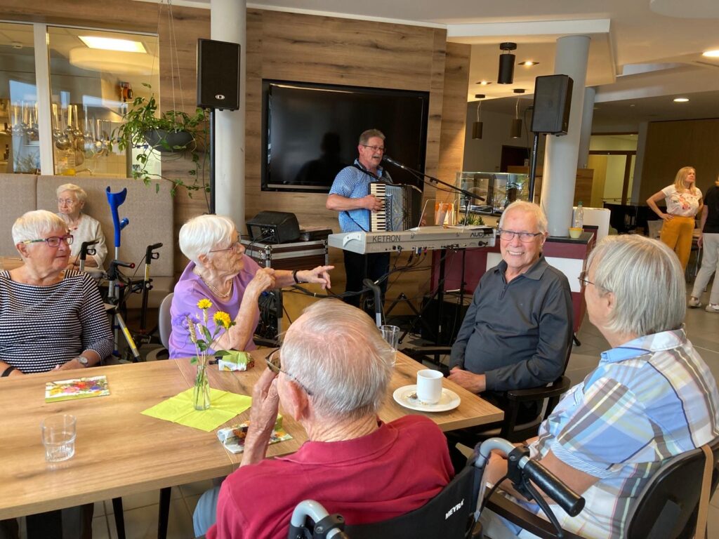 Eine Gruppe älterer Menschen sitzt lächelnd und plaudernd um einen Tisch in einem Gemeinschaftsraum, während ein Mann Akkordeon spielt und an einem Mikrofon im Hintergrund singt. Tassen, Teller und Blumen stehen auf dem Tisch.