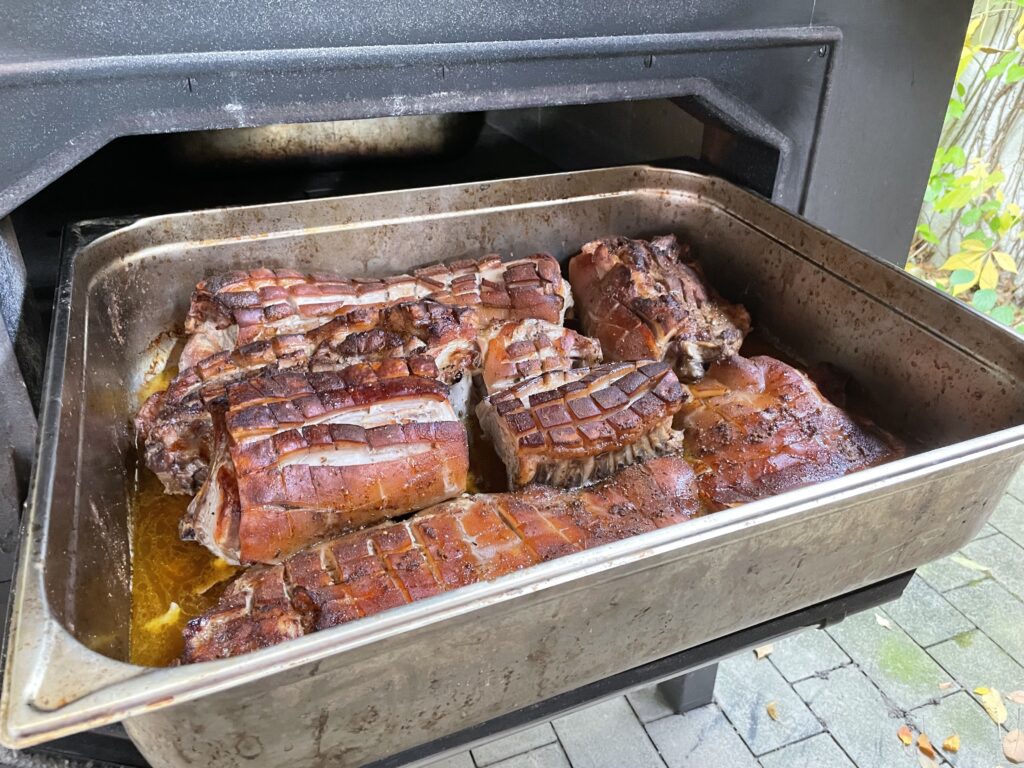 Eine Metallbratpfanne, gefüllt mit Schweinebratenstücken mit knusprigen, goldbraunen Grieben, steht in einem Ofen im Freien, und der Saft sammelt sich am Boden der Pfanne.