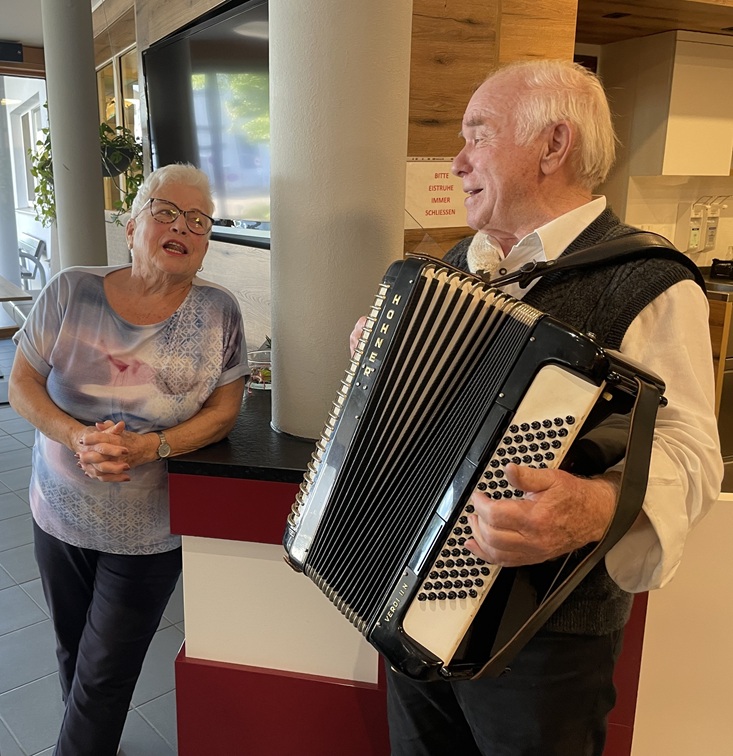 Ein älterer Mann spielt auf einem Akkordeon, während er lächelnd neben einer Frau steht, die sich an einen Tresen lehnt, ihn beobachtet und in einem gemütlichen Innenraum lächelt.