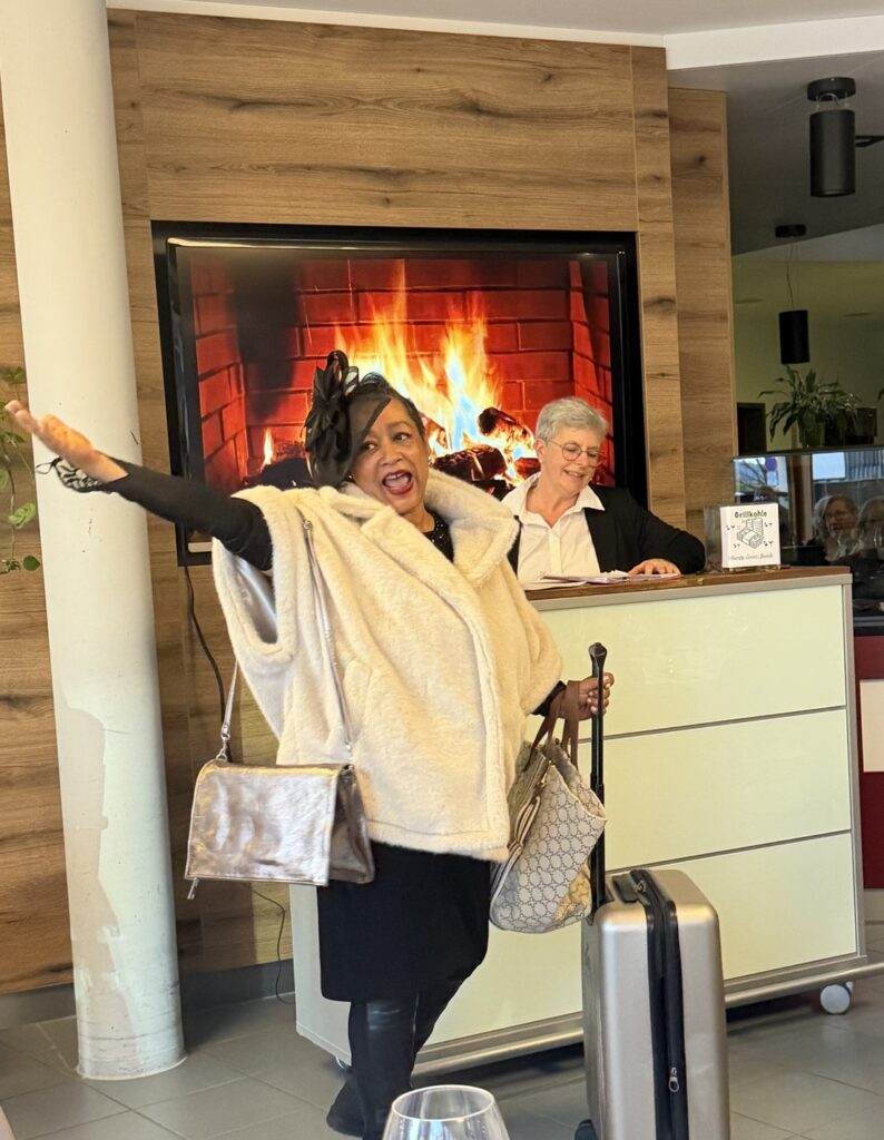 Eine Frau im Pelzmantel und mit Fascinator posiert fröhlich mit einem Koffer und einer Handtasche vor einem Empfangstresen. Eine lächelnde Empfangsdame steht hinter dem Tresen, und ein Bildschirm zeigt eine gemütliche Kaminszene.