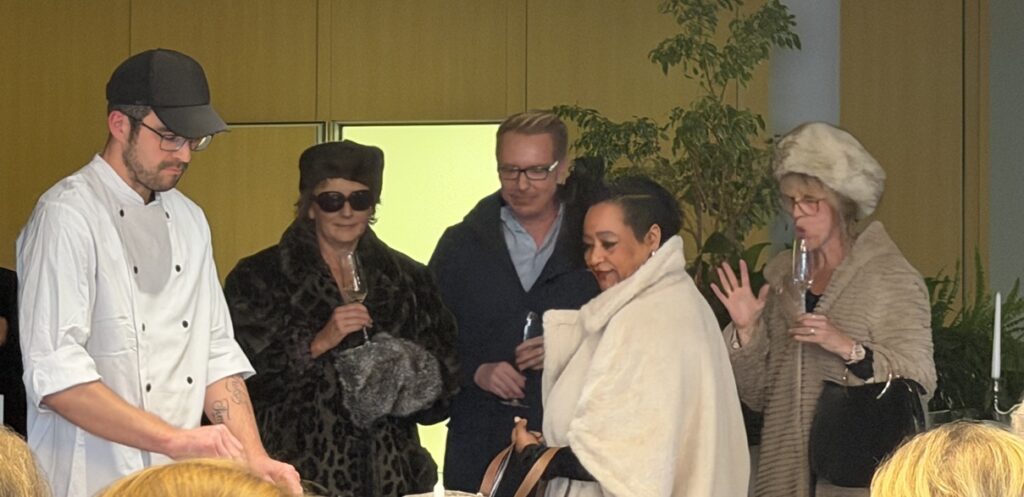 Ein Koch bereitet das Essen zu, während vier Personen in formeller Winterkleidung und mit Gläsern in der Hand in einem Innenraum mit Grünpflanzen im Hintergrund zusehen und sich unterhalten.