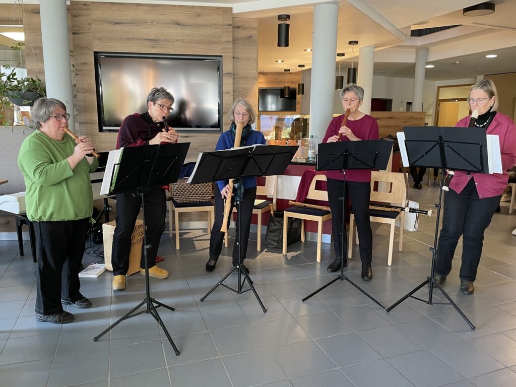Fünf ältere Erwachsene stehen in einem Raum und spielen Holzblasinstrumente, wobei jeder von ihnen die Notenblätter von den Ständern vor sich abliest. Sie spielen gemeinsam in einem modernen Raum mit Holzwänden und einem großen Fernseher im Hintergrund.