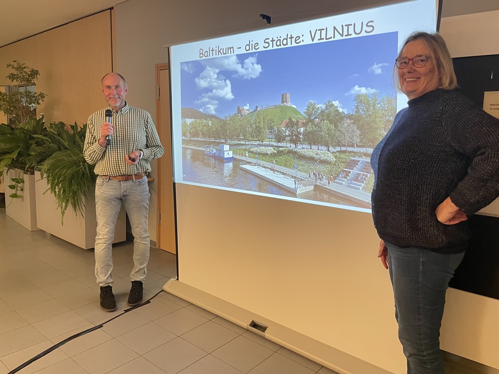 Ein Mann mit einem Mikrofon und eine Frau stehen vor einem projizierten Dia, das eine landschaftliche Ansicht von Vilnius, Litauen, zeigt. Das Dia trägt den Titel Baltikum - die Städte: VILNIUS. Sie scheinen einen Vortrag zu halten.
