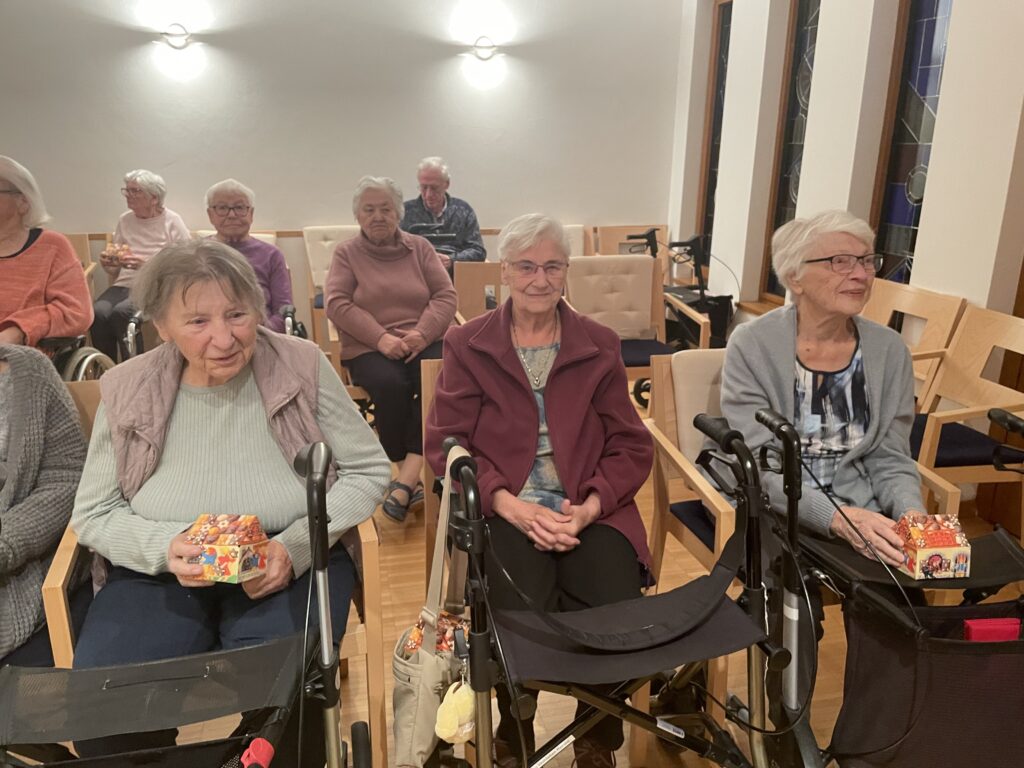 Eine Gruppe älterer Frauen sitzt in einem gut beleuchteten Raum, einige mit Gehhilfen, lächeln und halten kleine bunte Geschenkboxen in der Hand. Sie scheinen ein geselliges Beisammensein oder eine Veranstaltung zu genießen.