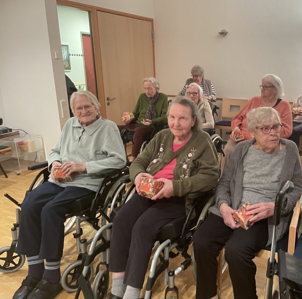Eine Gruppe älterer Frauen, einige in Rollstühlen, sitzt in einem Raum zusammen, hält kleine eingepackte Geschenke in der Hand und lächelt. Die Umgebung scheint ein Pflegeheim oder ein Gemeinschaftsraum mit Holzböden und hellen Wänden zu sein.