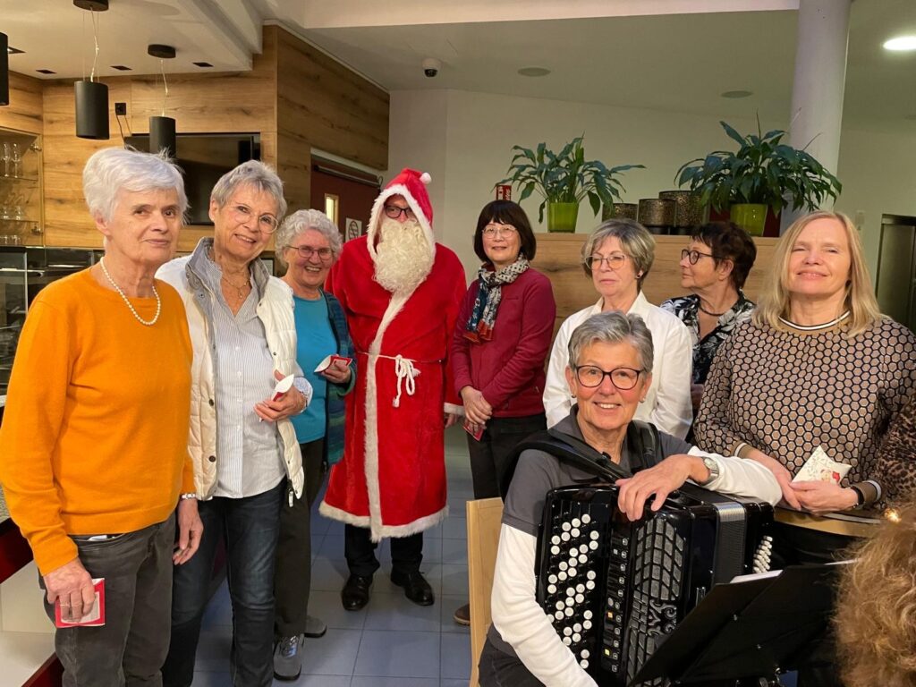 Eine Gruppe lächelnder älterer Erwachsener, von denen einer als Weihnachtsmann verkleidet ist, posiert gemeinsam in einem Haus. Eine Frau im Vordergrund hält ein Akkordeon, und im Hintergrund stehen Pflanzen auf einem Regal.