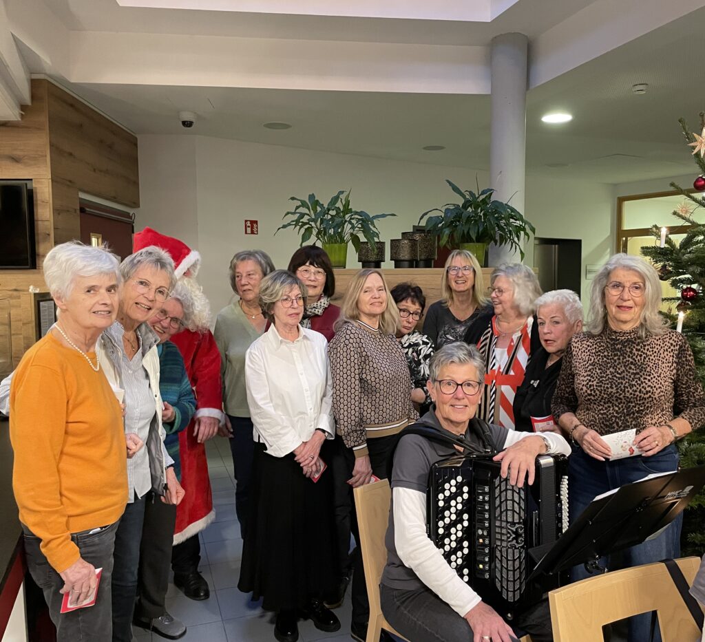 Eine Gruppe älterer Frauen, einige lächelnd, posiert in einem Haus in der Nähe eines Weihnachtsbaums. Eine Frau sitzt und spielt ein Akkordeon. Eine weitere Person ist als Weihnachtsmann verkleidet. Die Atmosphäre wirkt festlich und fröhlich.