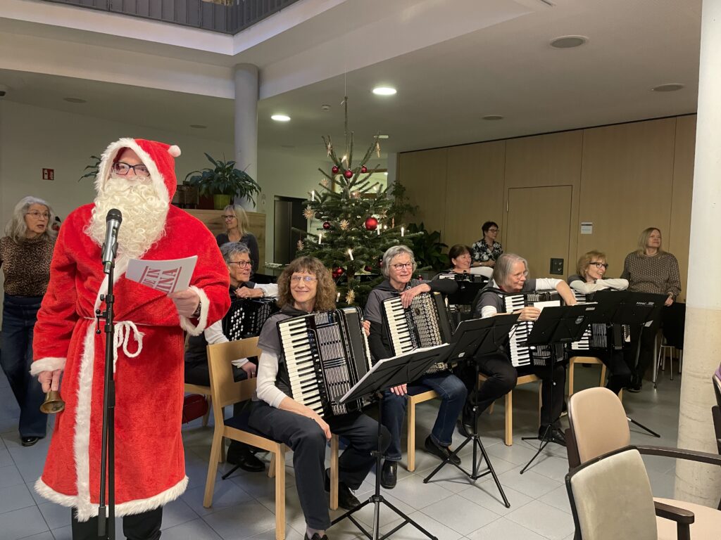 Eine als Weihnachtsmann verkleidete Person steht neben einer Gruppe von sitzenden Musikern, die in der Nähe eines geschmückten Weihnachtsbaums in einem hellen Innenraum Akkordeon spielen. Andere Personen stehen im Hintergrund und schauen zu.