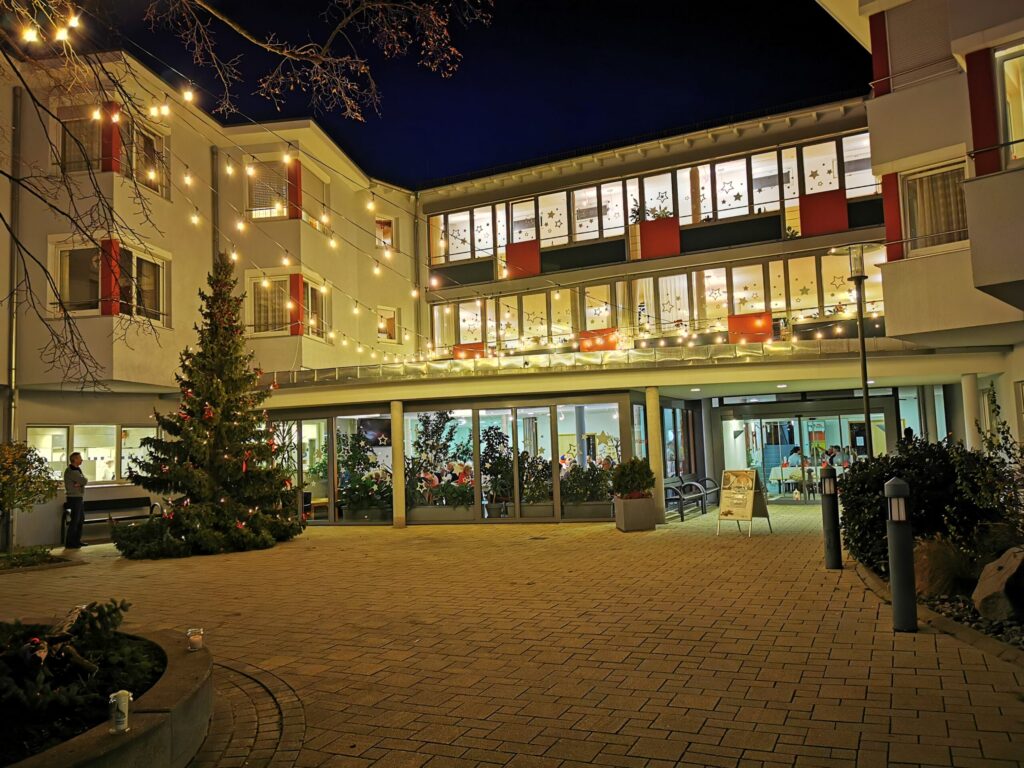 Ein mit Lichterketten und einem Weihnachtsbaum geschmückter Innenhof eines Gebäudes bei Nacht. Warmes Licht leuchtet aus den Fenstern, und in der hell erleuchteten Lobby sind Menschen zu sehen.