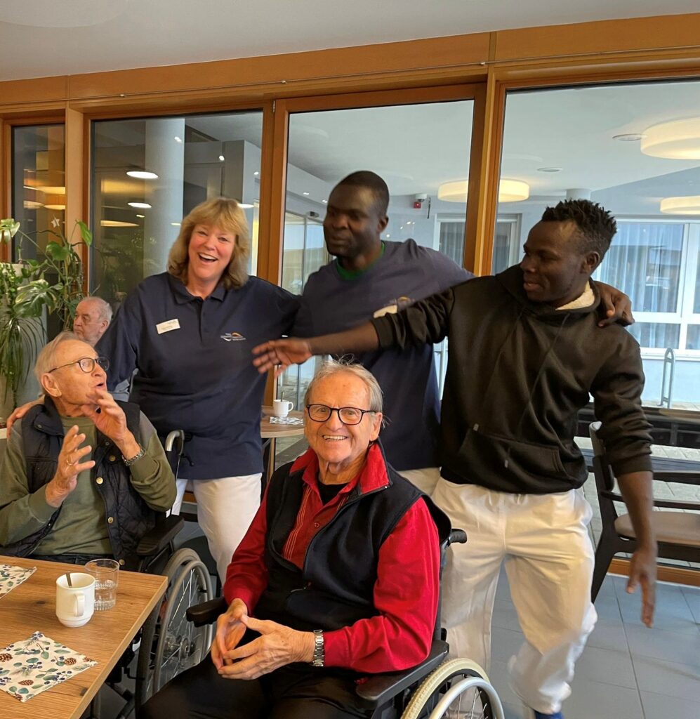 Zwei ältere Männer in Rollstühlen lächeln und unterhalten sich mit zwei stehenden Pflegern und einem weiteren stehenden Mann, die alle fröhlich wirken, in einem hellen Innenraum mit großen Fenstern.