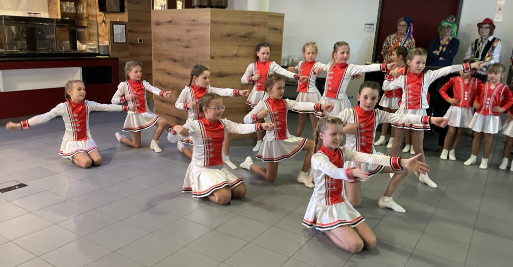 Eine Gruppe junger Mädchen in passenden rot-weißen Kostümen führt in einer Halle einen Synchrontanz auf, bei dem sie sich hinknien und ihre Arme ausstrecken, während einige Erwachsene im Hintergrund zusehen.