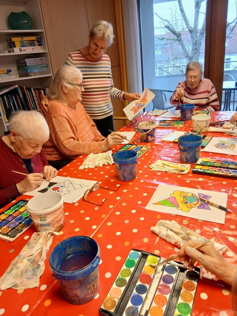 Eine Gruppe älterer Frauen sitzt um einen mit einem rot gepunkteten Tuch bedeckten Tisch und malt bunte Bilder mit Wasserfarben. Eine Frau steht da, beobachtet und plaudert, während die anderen an ihren Bildern arbeiten.