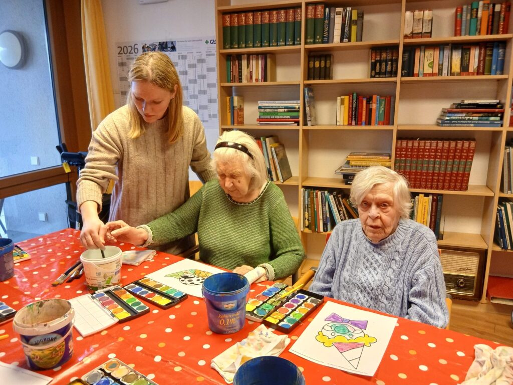 Eine junge Frau hilft zwei älteren Frauen bei der Aquarellmalerei an einem mit einem roten gepunkteten Tuch bedeckten Tisch. Kunstzubehör und fertige Bilder sind zu sehen; im Hintergrund stehen Bücherregale und ein Kalender.