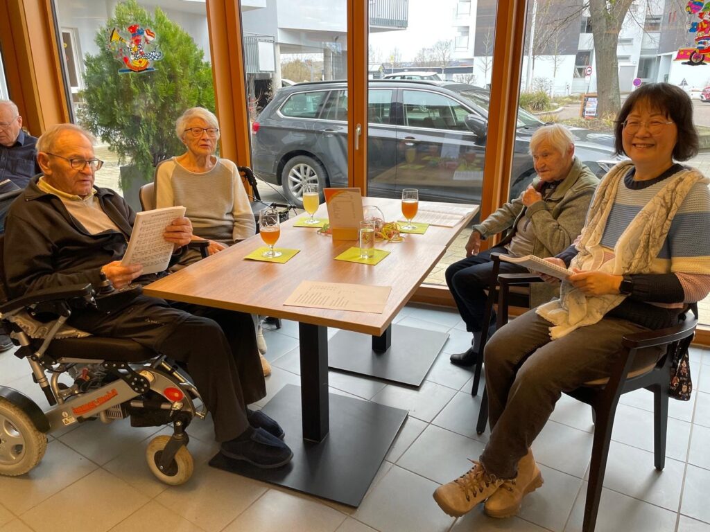Vier ältere Menschen sitzen um einen Tisch am Fenster, lächeln und halten Papiere in der Hand. Ein Mann sitzt in einem Rollstuhl, und auf dem Tisch stehen Getränke. Draußen ist ein Auto in der Nähe von Bäumen und Gebäuden geparkt.