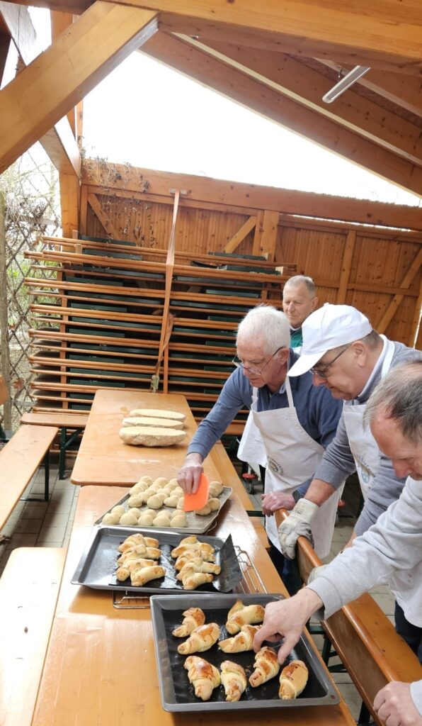 In einem hölzernen Pavillon im Freien bereiten vier ältere Männer in Schürzen frisch gebackene Brötchen auf Tabletts vor, während auf den Tischen daneben Teigkugeln und Brotlaibe liegen.