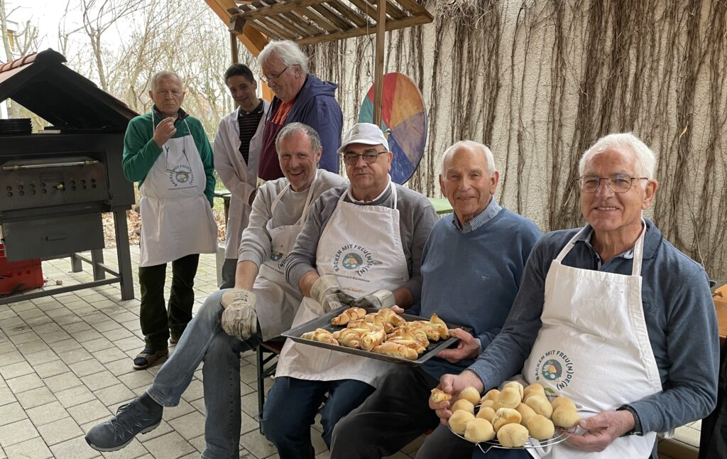 Sieben lächelnde ältere Menschen, einige mit Schürzen und Handschuhen, posieren im Freien vor einem Holzofen. Zwei halten Tabletts mit frisch gebackenen Brötchen und Gebäck. Zweige bedecken die Wand im Hintergrund.