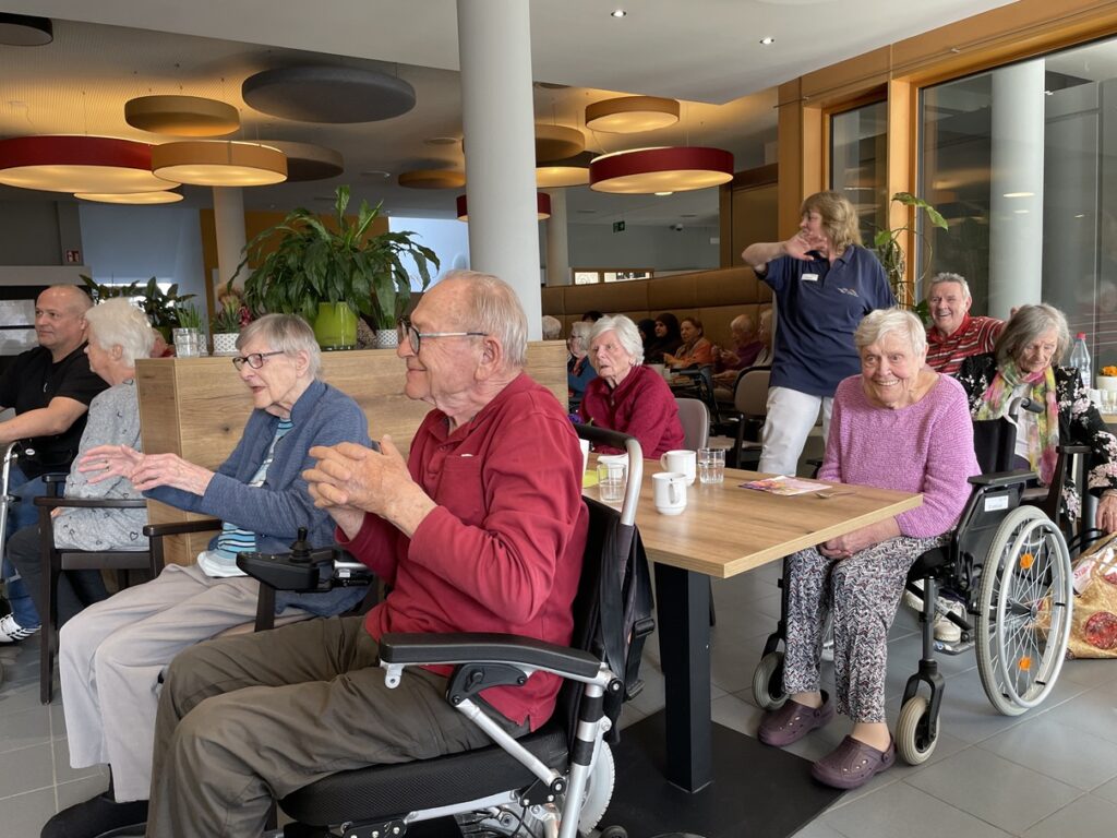 Eine Gruppe älterer Menschen, einige in Rollstühlen, sitzt in einem hell erleuchteten Café oder Lounge-Bereich zusammen, lächelt und genießt die Gesellschaft der anderen. Eine Betreuungsperson steht in der Nähe, und von der Decke hängen bunte Lichter.