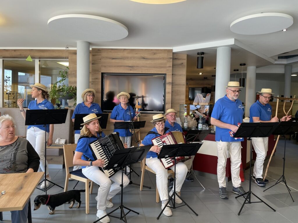 Eine Gruppe älterer Erwachsener in blauen Hemden, weißen Hosen und Strohhüten spielt Akkordeon und singt in einem modernen Innenraum und unterhält sitzende Menschen. Ein kleiner schwarzer Hund liegt auf dem Boden im Vordergrund.