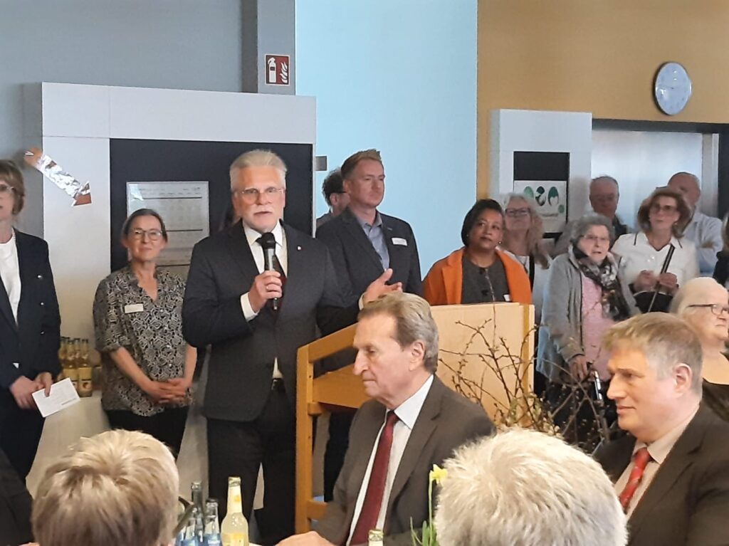 Ein Mann im Anzug spricht bei einer formellen Veranstaltung in einem Haus in ein Mikrofon, während andere um ihn herum stehen und sitzen und aufmerksam zuhören. Auf den Tischen stehen Getränke, und die Leute scheinen in die Veranstaltung vertieft zu sein.