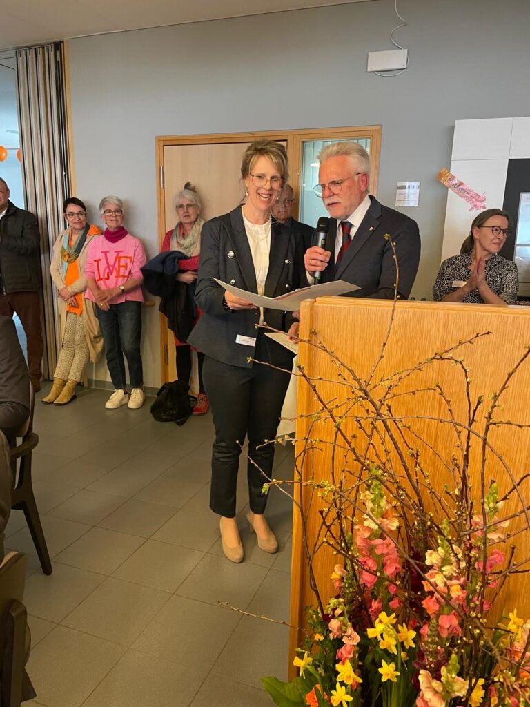 Ein Mann spricht in ein Mikrofon an einem Podium, neben ihm eine Frau, beide lächeln. Mehrere Personen stehen im Hintergrund, und ein Blumengesteck schmückt die Vorderseite des Podiums.