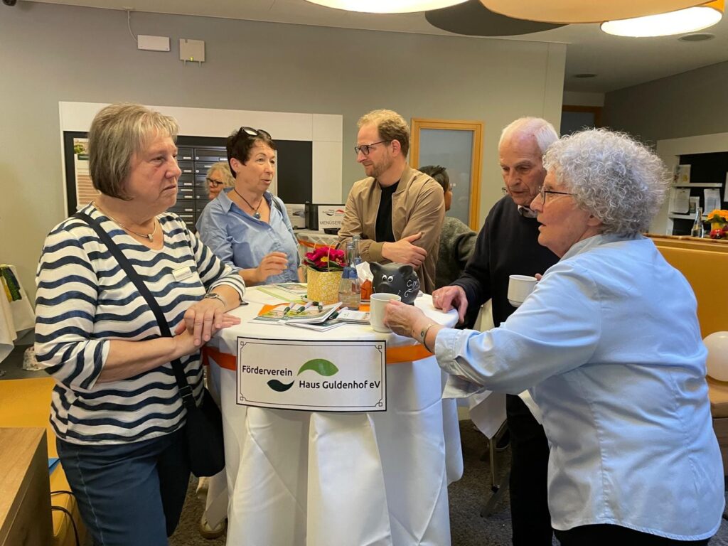 Fünf Personen, drei Frauen und zwei Männer, stehen um einen hohen Tisch mit Broschüren und Blumen. Sie scheinen sich bei einer Indoor-Veranstaltung zu unterhalten. Auf dem Tisch steht ein Schild mit der Aufschrift Förderverein Haus Guldenhof e.V.