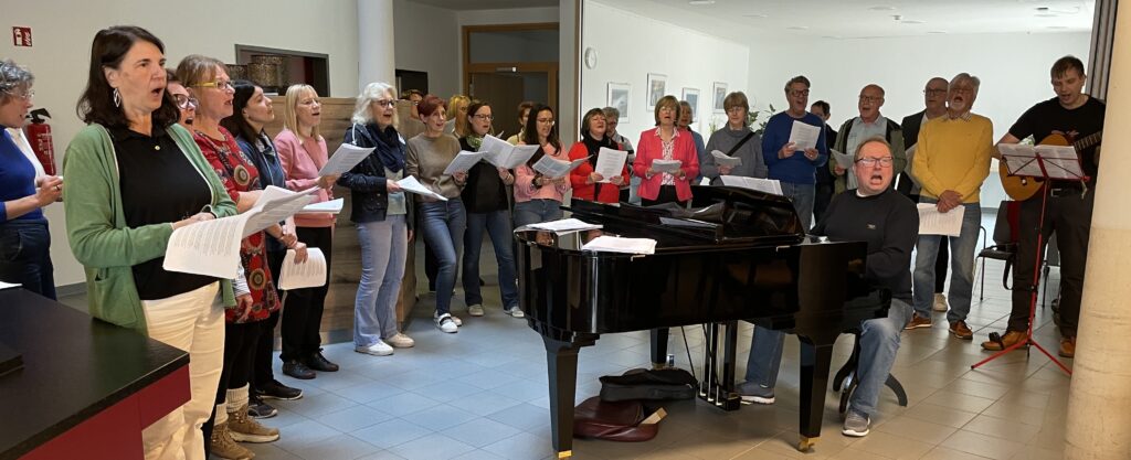 Eine Gruppe Erwachsener steht im Halbkreis um einen schwarzen Flügel und singt von Notenblättern, während ein Mann Klavier und ein anderer Gitarre in einem hellen Innenraum spielt.