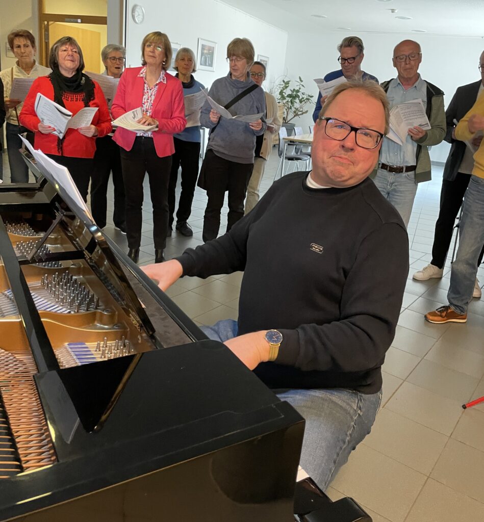 Ein Mann mit Brille spielt auf einem Flügel und blickt dabei leicht zur Seite, während eine Gruppe von Menschen, die hinter ihm steht, in einem hellen Innenraum von Notenblättern singt.