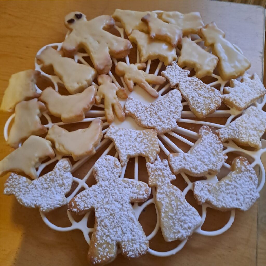 Auf einem weißen Kühlregal auf einer hölzernen Unterlage sind verschiedene Kekse aus Zuckerguss und Puderzucker in Form von Menschen, Katzen und Vögeln angeordnet.