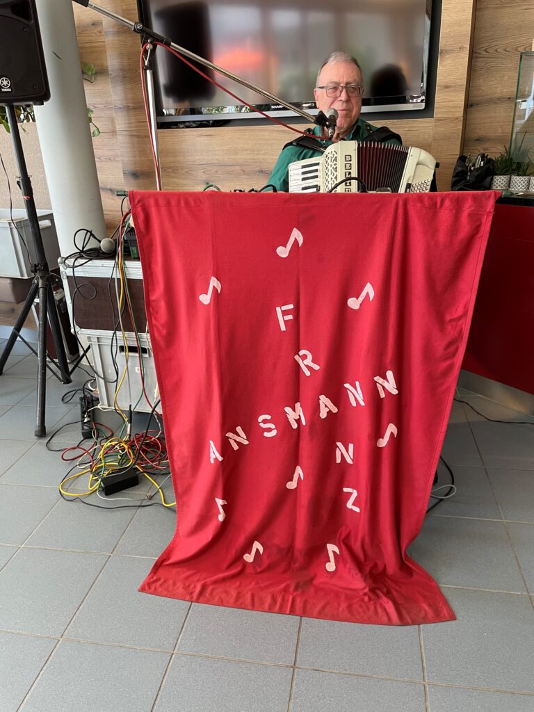 Ein älterer Mann spielt ein Akkordeon und singt in ein Mikrofon hinter einem roten Tuch, das mit Musiknoten und den Buchstaben FR ANSMANN verziert ist. Auf dem gefliesten Boden sind Musikgeräte und bunte Kabel zu sehen.