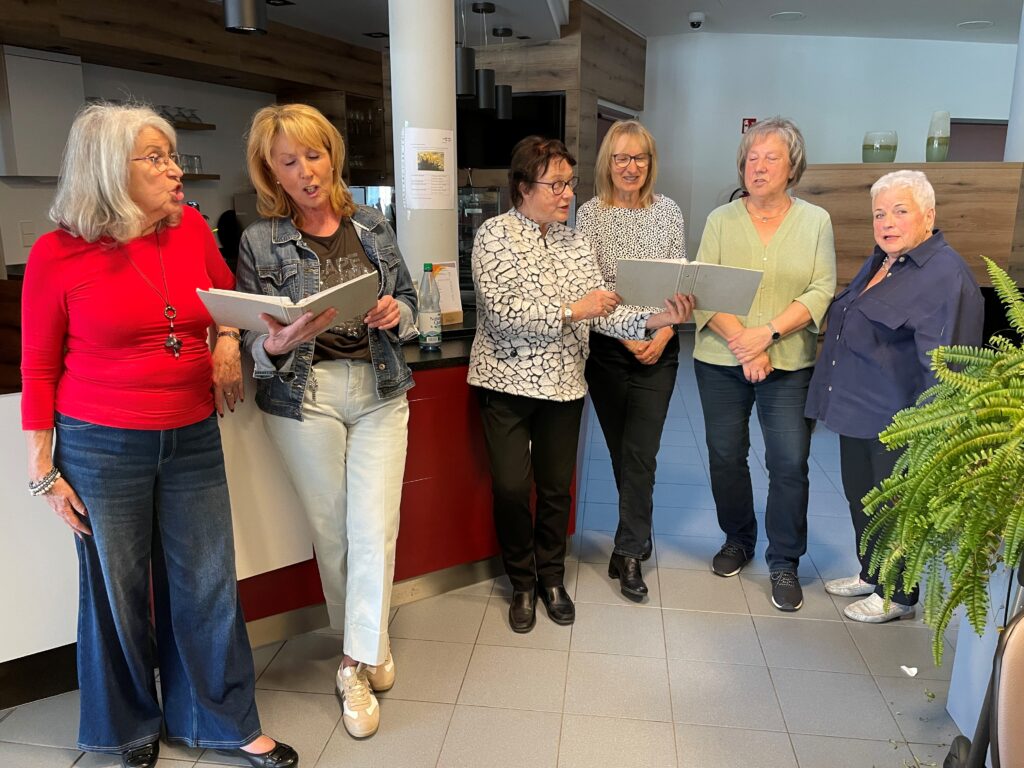 Sechs ältere Frauen stehen in einem Innenraum zusammen, vier von ihnen halten ein Blatt Papier in der Hand und lesen daraus vor. Sie scheinen zu singen oder zu rezitieren und sind leger gekleidet. Der Schauplatz ist eine moderne Küche oder ein Gemeinschaftsraum.